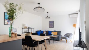 Una sala de estar moderna con una mesa de comedor de madera, sillas negras, un sofá gris con cojines azules y naranjas, y un sillón gris. Hay un televisor sobre un mueble negro, un florero con flores sobre la mesa y un cuadro enmarcado en la pared.