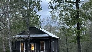 A rustic cabin with a porch, surrounded by trees and a grassy slope.