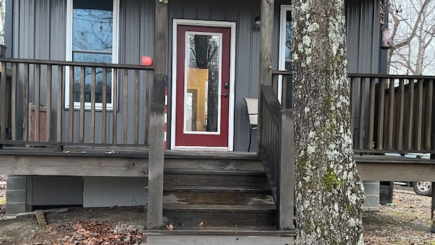A wooden cabin with a front porch, a red door, and a stone foundation.