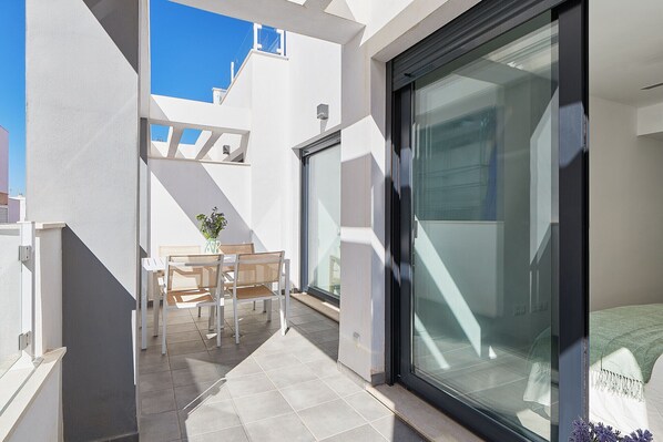 A modern outdoor balcony with a glass door, a wooden table, and a potted plant.