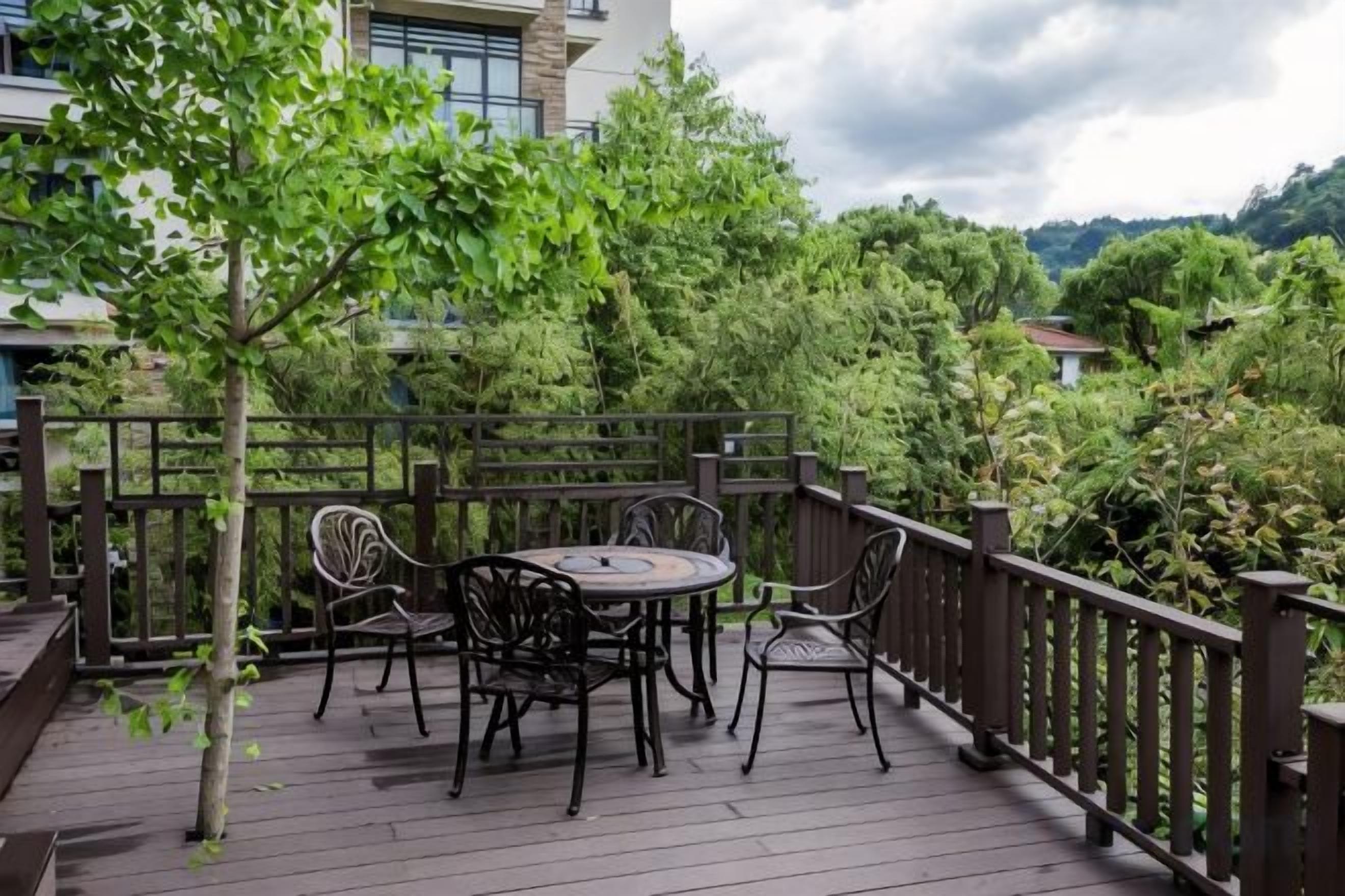 A wooden deck with a round table and chairs, surrounded by a wooden railing, overlooking a lush green landscape.