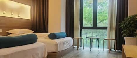 A hotel room with a large window, a bed with white bedding and blue pillows, a small table, and a chair.
