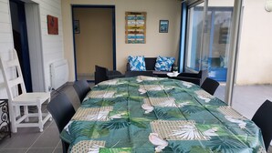 Uma área de refeições com uma grande mesa coberta por uma toalha de folha tropical, rodeada por cadeiras pretas. Há também um espaço de estar com um sofá e uma mesa de centro.