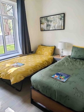 Ein Zimmer mit zwei Betten, einem Fenster mit Vorhängen, einem Bild an der Wand und einer Lampe auf dem Nachttisch.