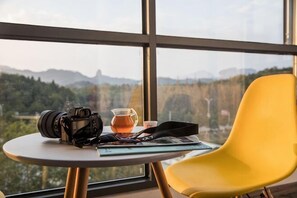 Een tafel met een camera, een kop thee en een tijdschrift, een gele stoel, en uitzicht op bergen door grote ramen.