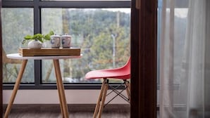 A window with a view of trees, a table with a plant and teapots, and a red chair.