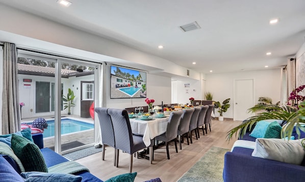 A modern dining area with a pool view, a dining table set with plates, glasses, and flowers, and a TV screen displaying a pool scene. - Heated Pool~Stellar stay in 7BD villa~Playground (Fort Lauderdale)