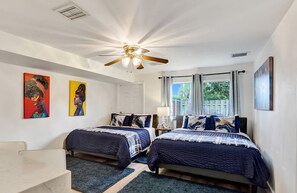 A bedroom with two beds, a ceiling fan, artwork on the walls, and a window with curtains.