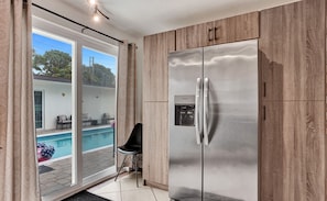 A modern kitchen with a stainless steel refrigerator, wooden cabinets, and a sliding glass door leading to a pool area. - Heated Pool~Stellar stay in 7BD villa~Playground (Fort Lauderdale)