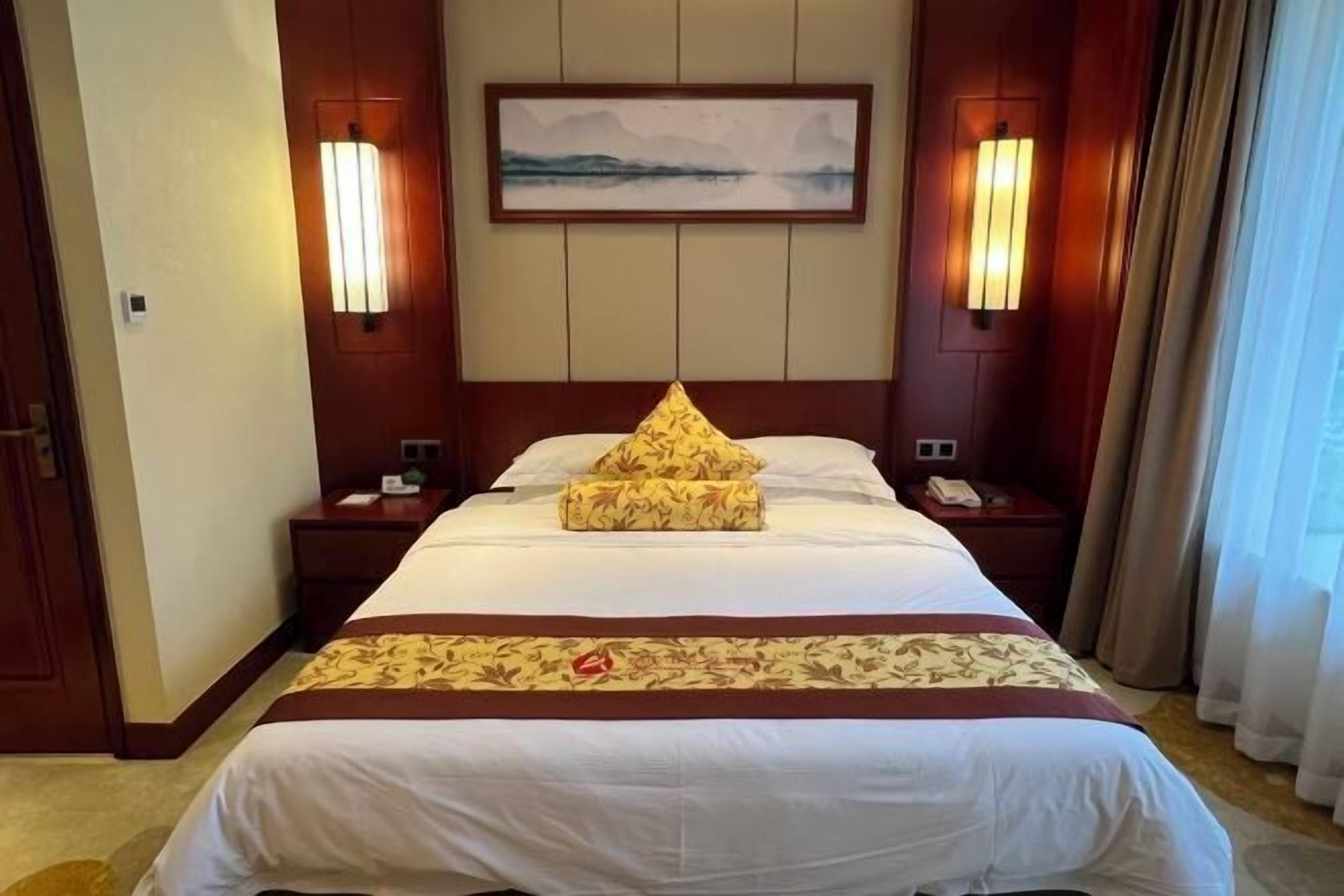 A hotel room with a large bed, two bedside tables, a nightstand, a wall-mounted lamp, and a painting above the bed.