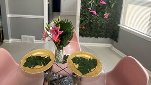 Un comedor con sillas rosadas, una mesa de vidrio con centros de mesa de pan de oro y un jarrón con flores rosas. En la pared, un fondo de hojas verdes y flamencos rosados.
