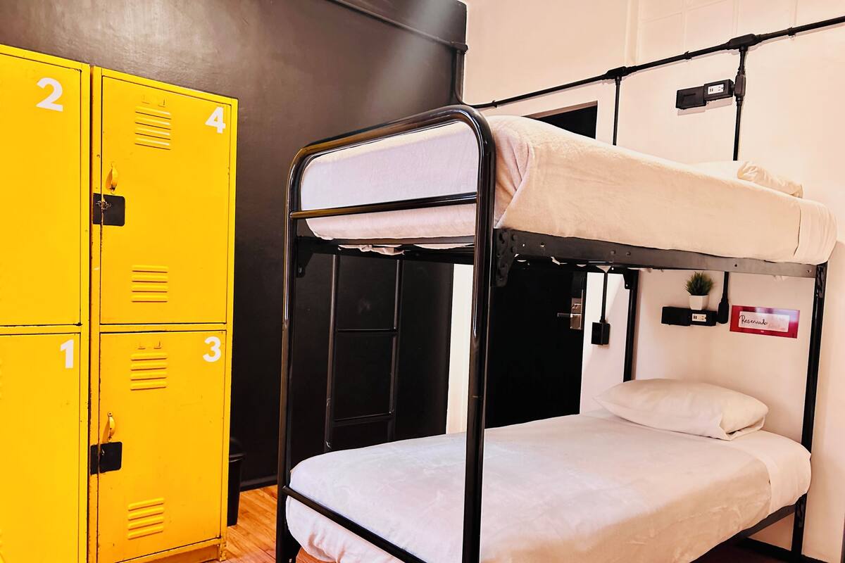 Ein Etagenbett mit weißer Matratze und einem Einzelbett darunter, ein gelbes Längsschrank-System und eine kleine Pflanze auf einem Regal.