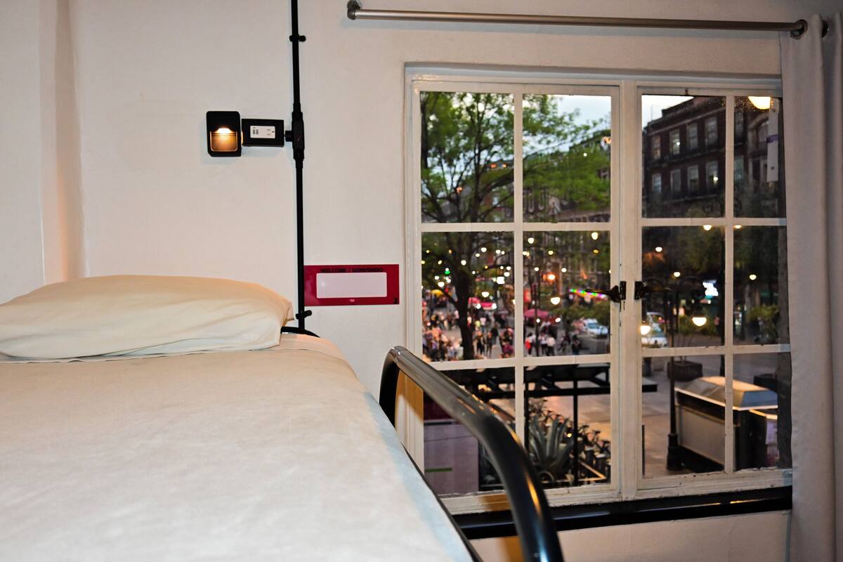 Ein Hotelzimmer mit Bett, Nachttisch, Wandlampe und einem großen Fenster mit Blick auf die Straße.