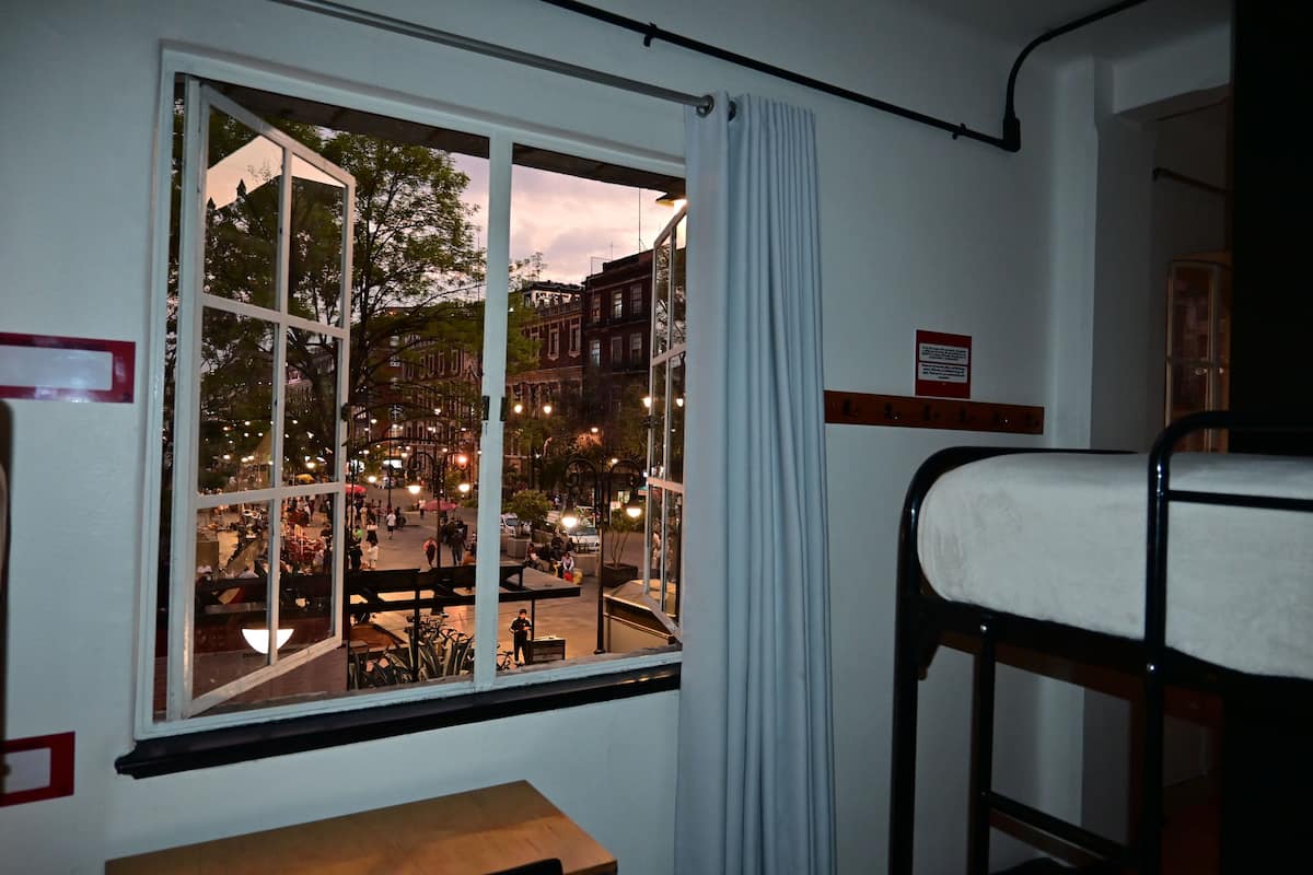 Ein Zimmer mit großem Fenster mit Blick auf eine belebte Straße in der Dämmerung, einem Etagenbett und einem kleinen Tisch.
