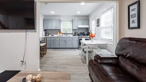 Un salon moderne avec un canapé en cuir marron, un téléviseur fixé au mur et un coin cuisine doté de meubles gris et d’une table à manger.