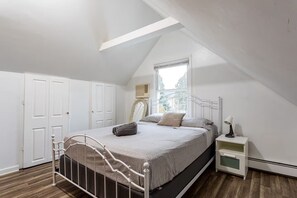 Une chambre avec un plafond en pente, un lit avec un cadre en métal, une table de chevet avec une lampe et une fenêtre donnant sur les arbres.