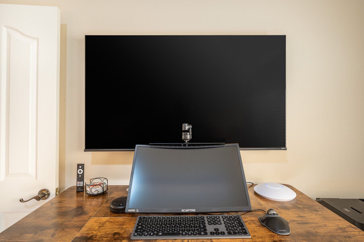 A room with a large flat-screen TV mounted on the wall, a laptop on a wooden desk, a keyboard, a mouse, and a remote control.