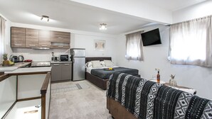 Ein kompaktes Hotelzimmer mit Küchenzeile, einem Bett mit gemustertem Überwurf, einem kleinen Tisch mit einer Ananas und einer Flasche sowie einem an der Wand montierten Fernseher.
