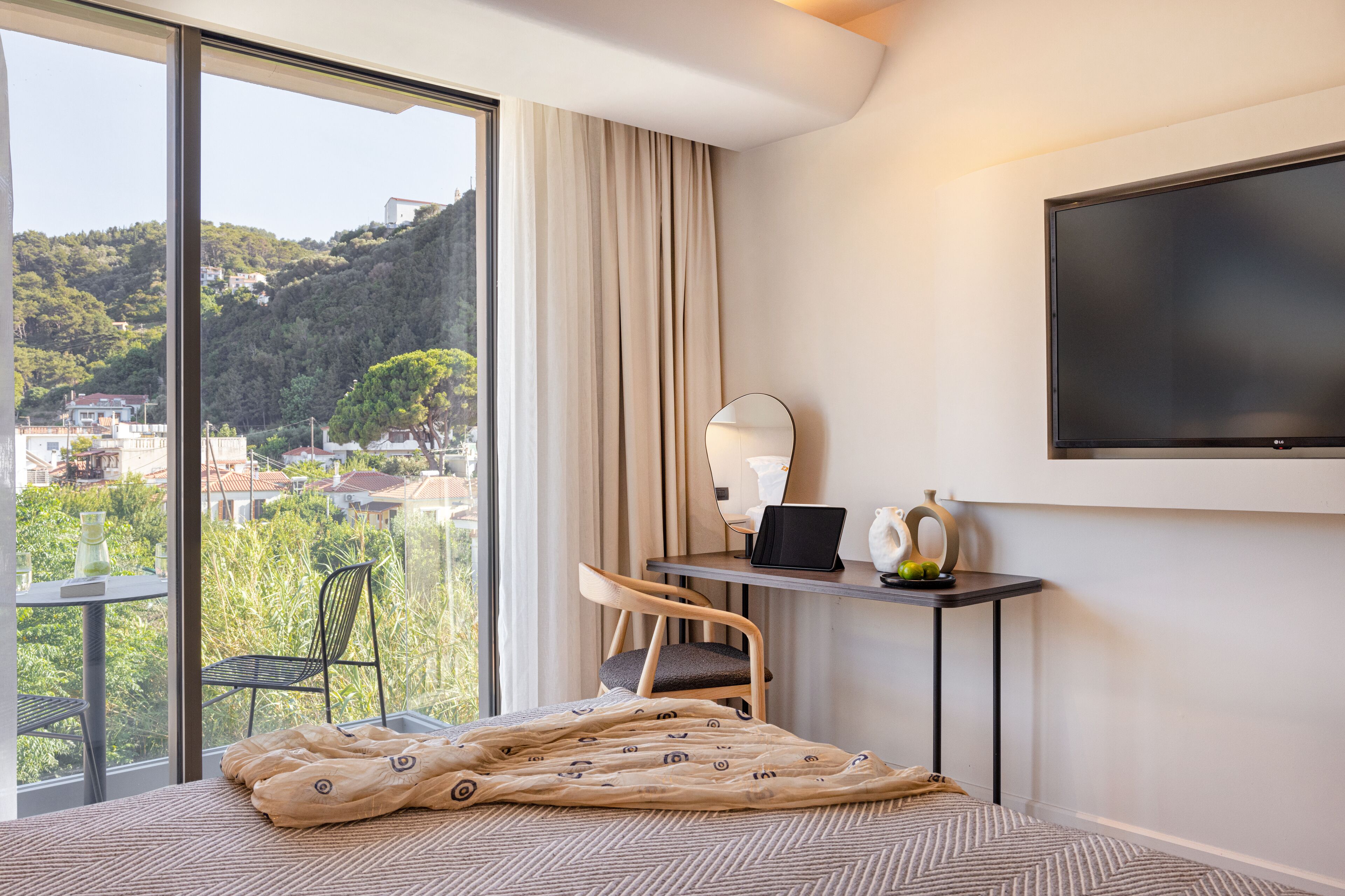 Ein Hotelzimmer mit großem Fenster, einem Bett mit gemustertem Überwurf, einem Schreibtisch mit Laptop, einem Sessel und einem an der Wand befestigten Flachbildfernseher.