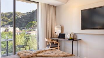 Ein Hotelzimmer mit großem Fenster, einem Bett mit gemustertem Überwurf, einem Schreibtisch mit Laptop, einem Sessel und einem an der Wand befestigten Flachbildfernseher.