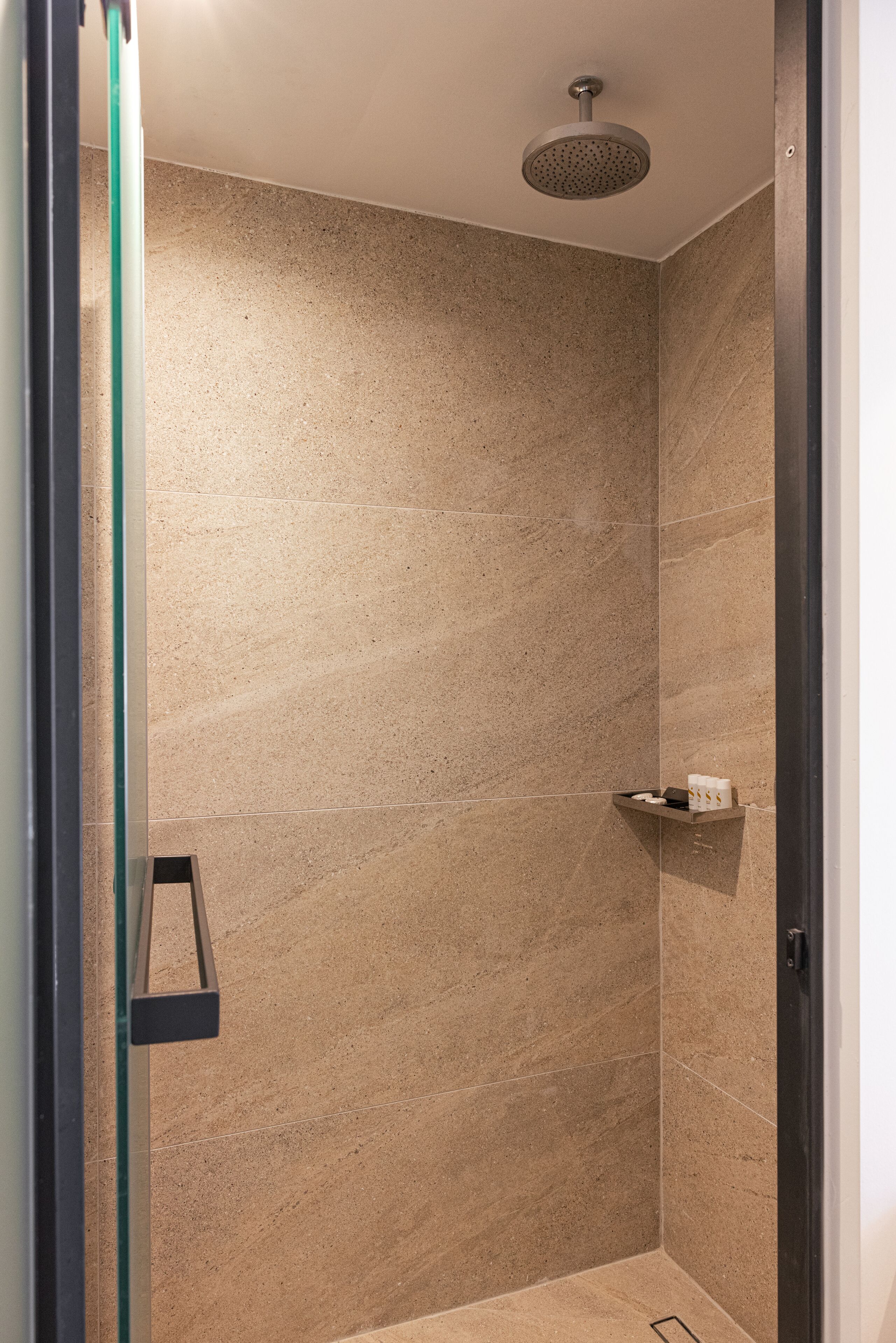 A modern shower with a rain showerhead, beige marble walls, and a small shelf with toiletries.