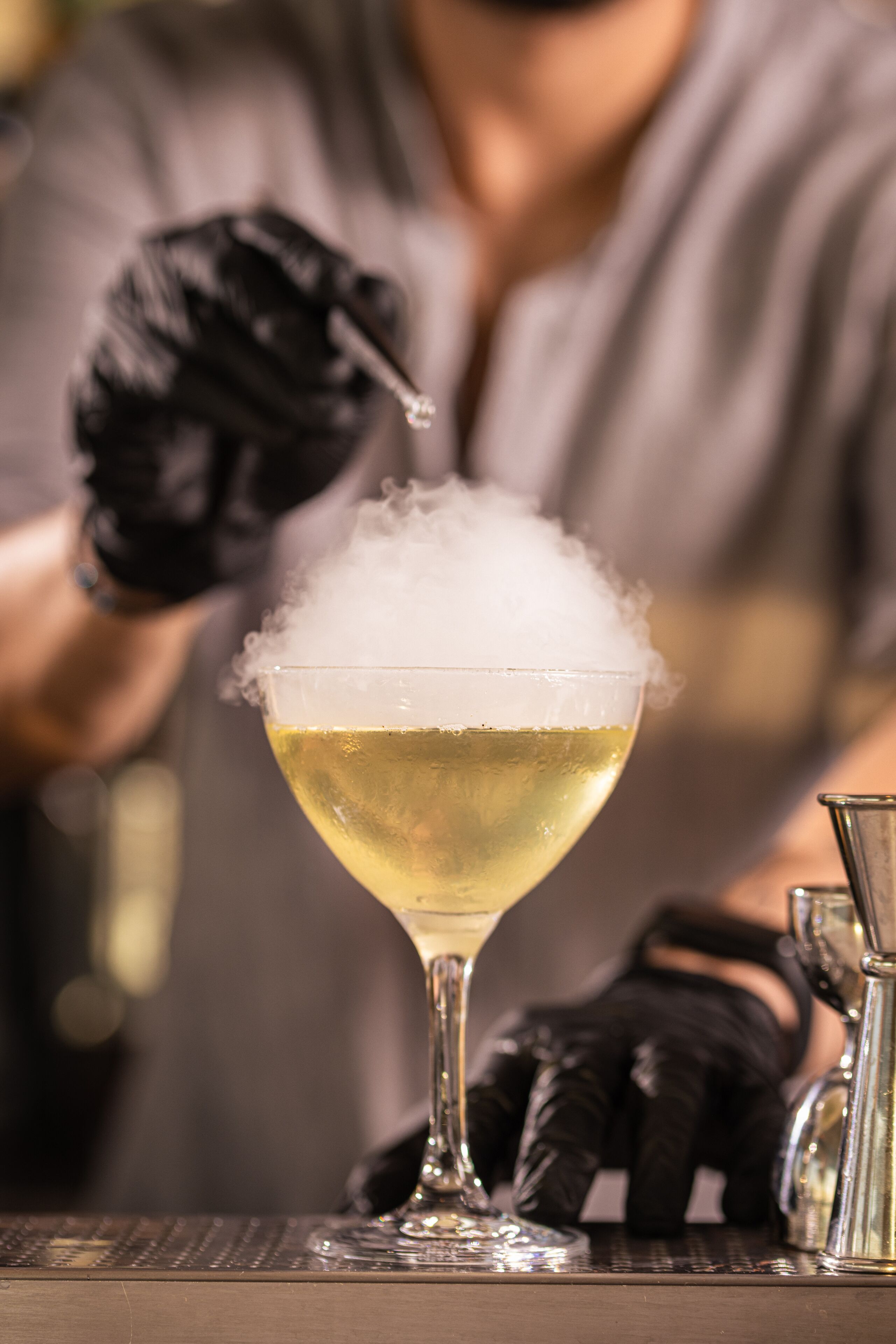 A cocktail with a foamy top, a clear glass, and a person wearing black gloves.