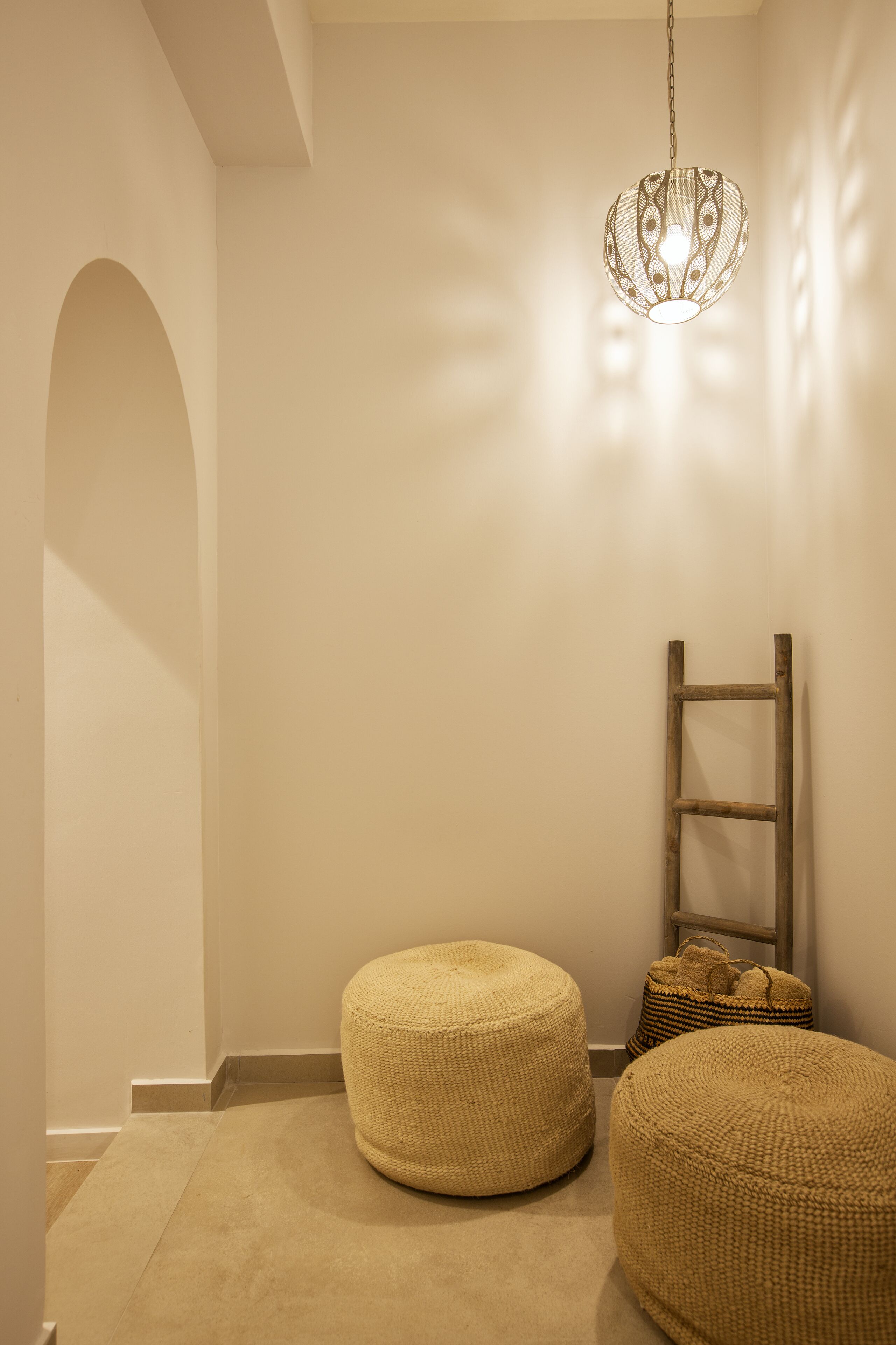 Una habitación con un colgador de luz, dos otomanas tejidas, una escalera de madera y una pequeña cesta tejida frente a una pared lisa.