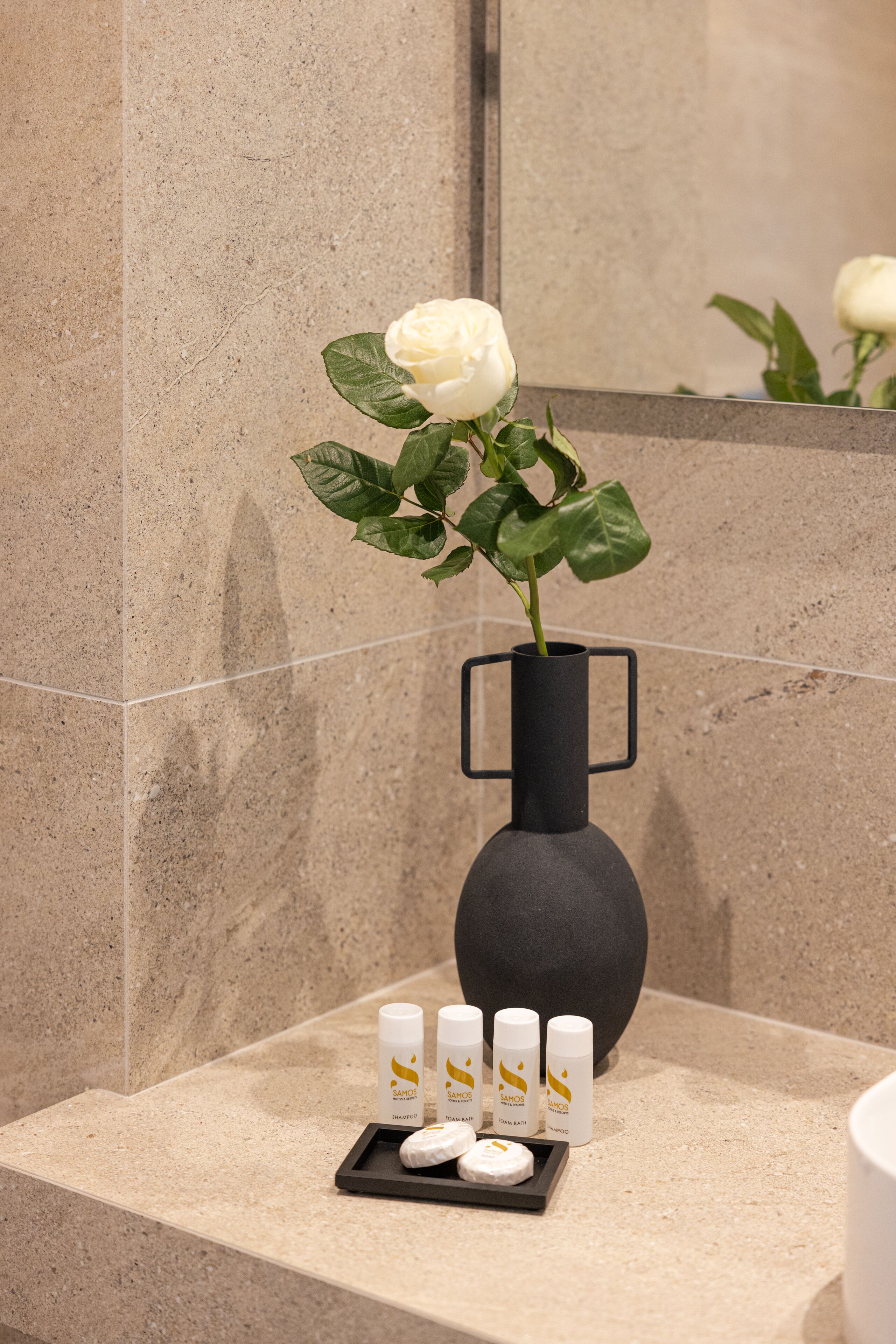 A bathroom with a marble countertop, a black vase with a white rose, and toiletries.