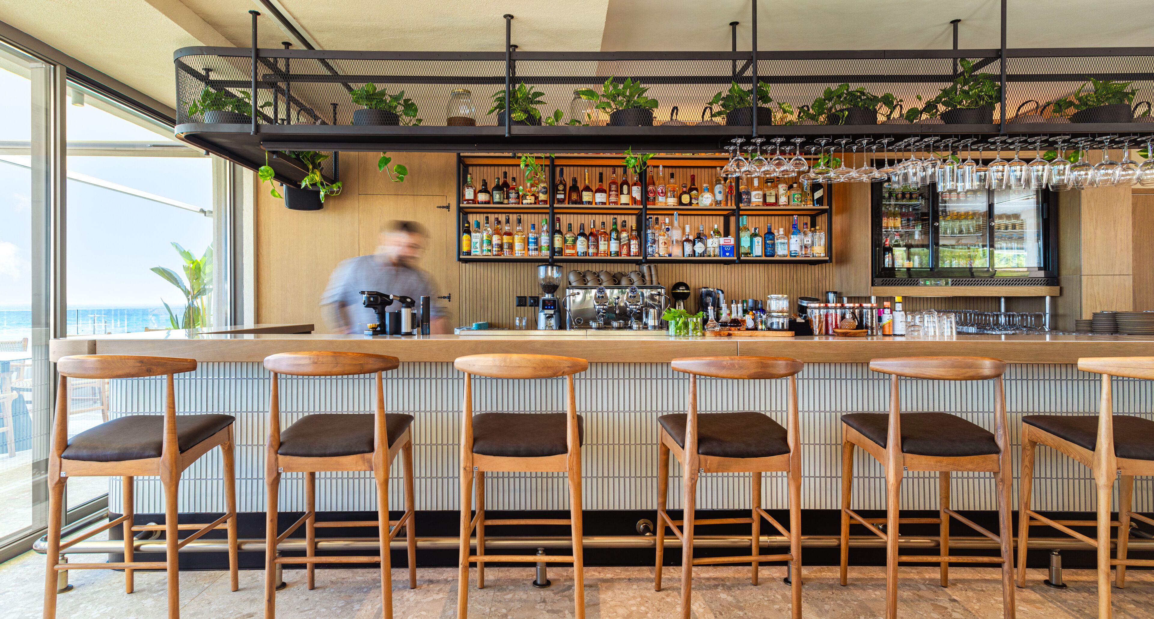 A bar area with wooden stools, a bar counter, shelves with bottles, and hanging glasses.