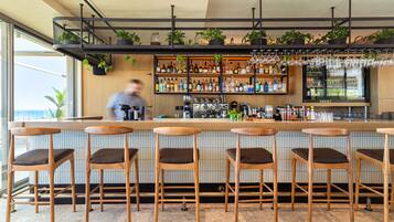 Una zona de bar con taburetes de madera, una barra, estantes con botellas y copas colgantes.