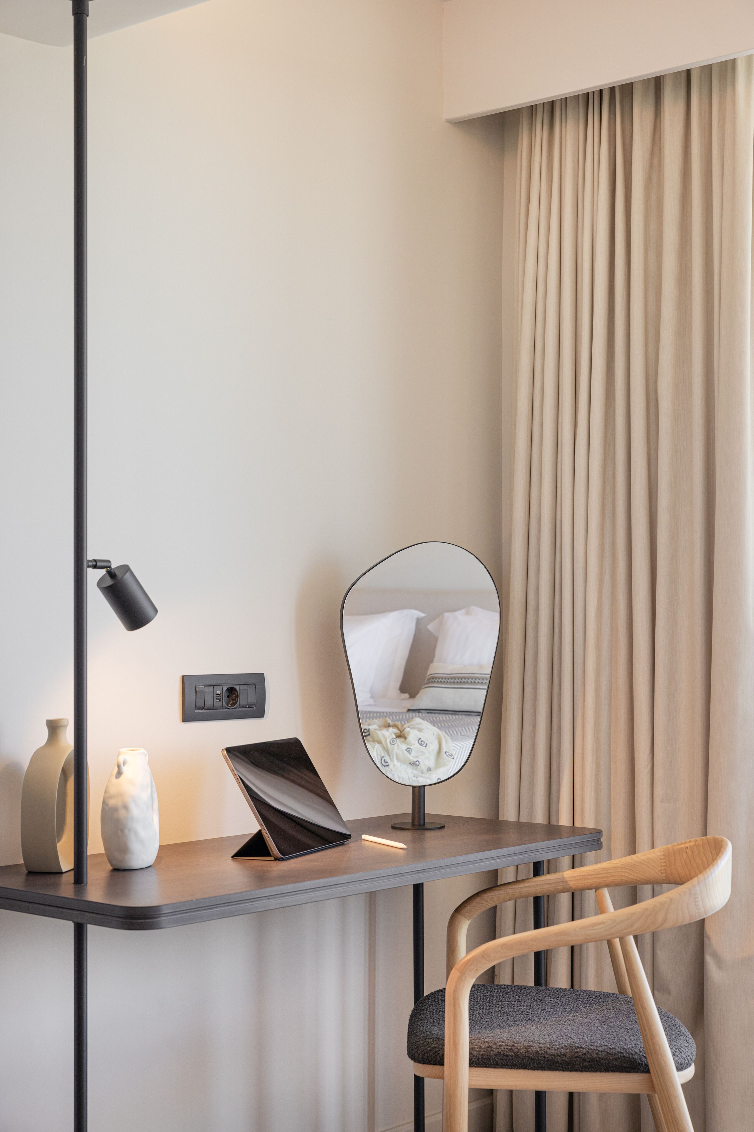 Una habitación de hotel moderna con escritorio, silla, lámpara, espejo y un ventanal con cortinas.