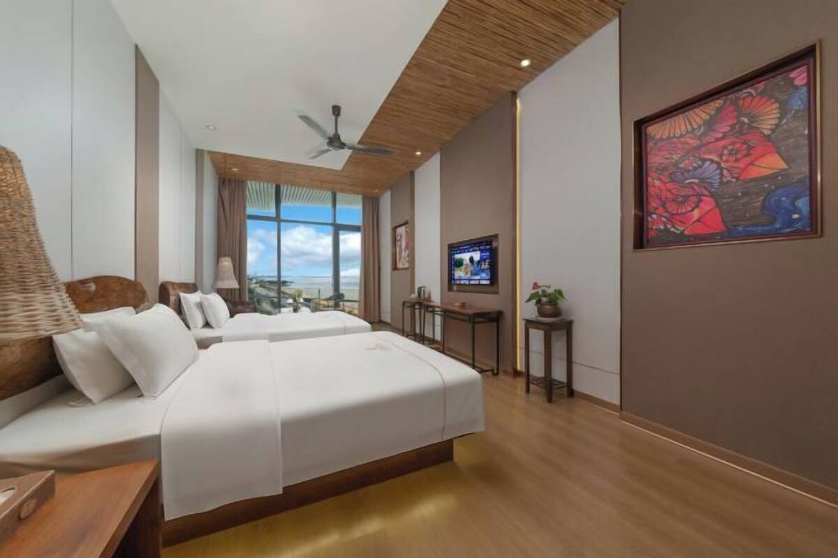 Habitación de hotel con cama, televisión, ventilador de techo, ventana con vistas y un cuadro en la pared.