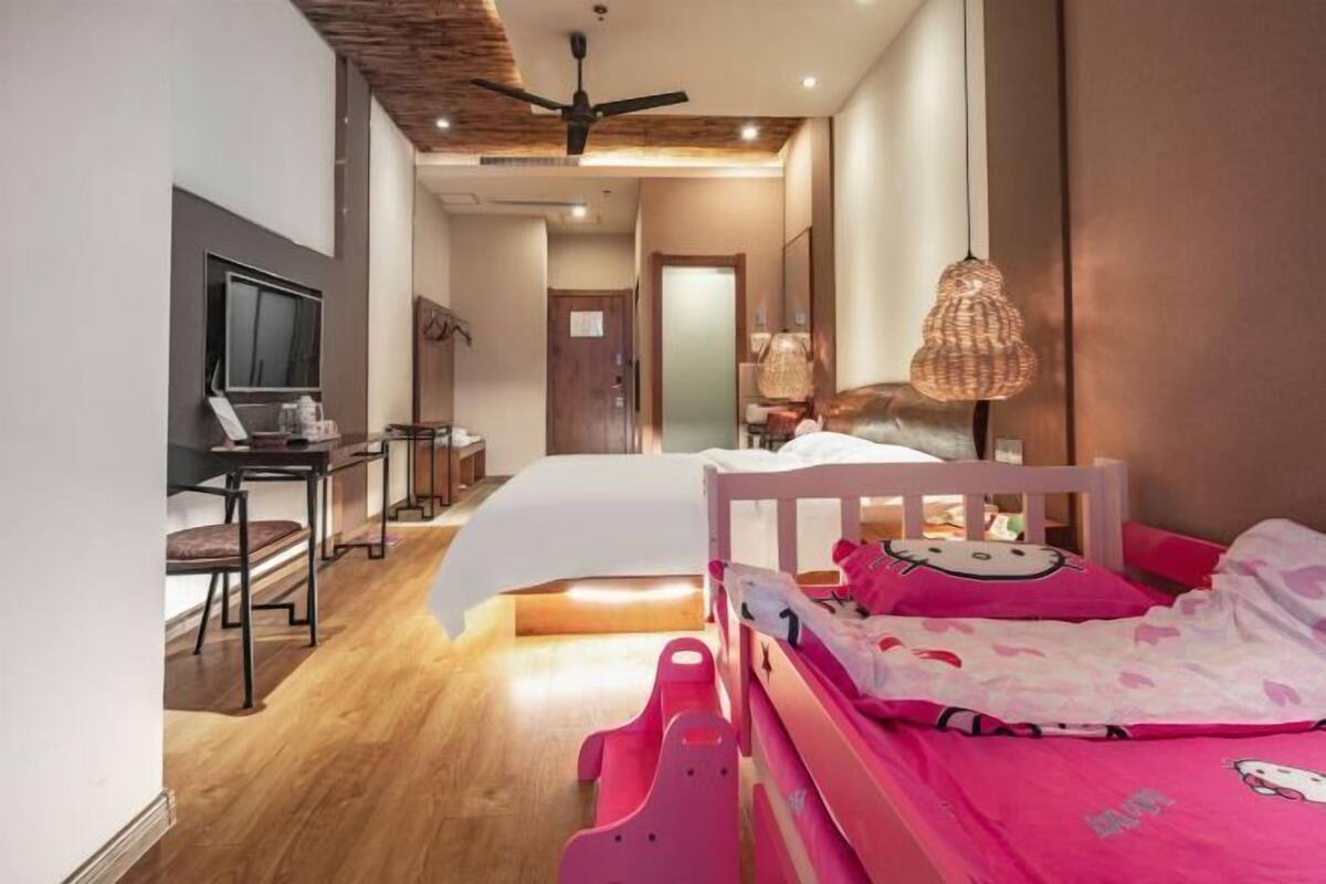 Una habitación de hotel moderna con piso de madera, una cama con ropa de cama rosa, un escritorio con una silla, un televisor y un ventilador de techo.