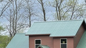 A two-story house with a green metal roof, a front porch with wooden stairs, and a small front yard with a lawn and shrubs. - Relax and Unwind on the Caddo River in Beautiful Arkansas! Book now! (Amity)