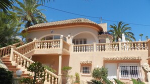 Uma casa de dois andares com varanda, escadaria e um jardim com palmeiras.