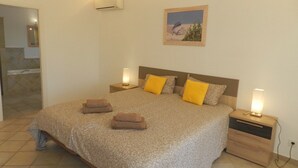 Quarto com uma cama grande, duas mesas de cabeceira, um criado-mudo, um aparelho de ar condicionado, um quadro na parede e um banheiro visível por uma porta aberta.