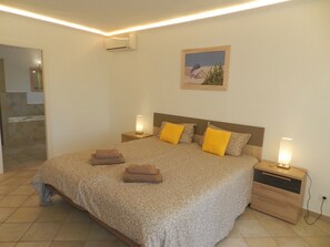 A bedroom with a large bed, two bedside tables, a nightstand, an air conditioning unit, a framed picture on the wall, and a bathroom visible through an open door. - Villa Ambiente para 4 personas (Calpe)
