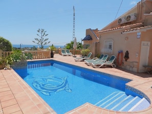 Ein Swimmingpool mit Delphin-Motiv, umgeben von Liegestühlen und Blick aufs Meer.
