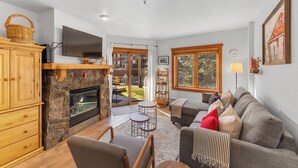 A cozy living room with a stone fireplace, wooden furniture, and large windows offering a view of the outdoors. - Red Hawk Lodge #2213 by Summit County Mountain Retreats (Keystone)