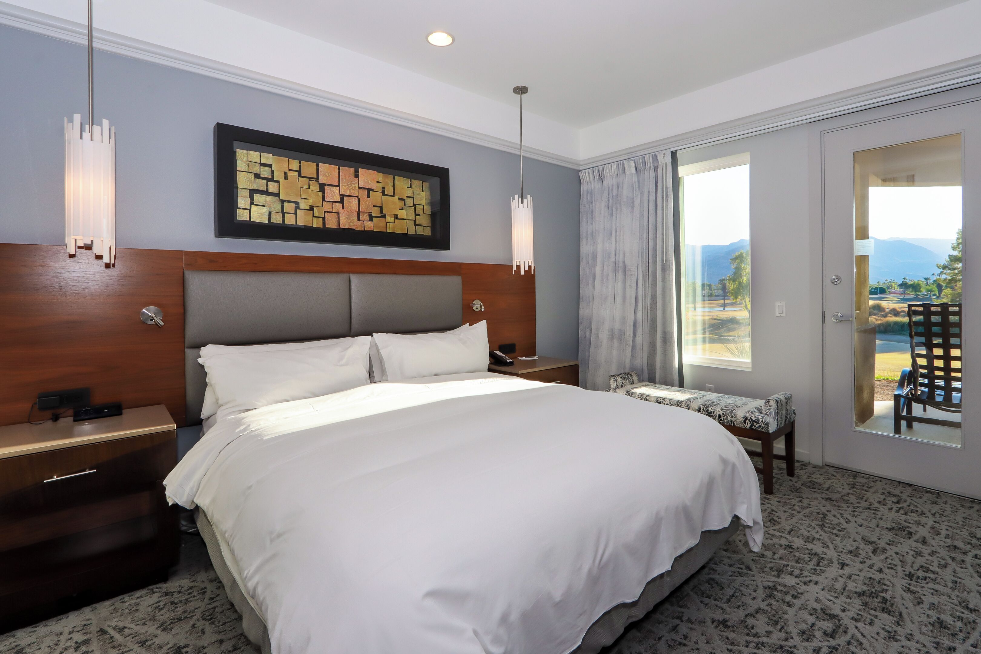 Habitación de hotel con una cama grande, cabecera de madera, mesita de noche, cuadro enmarcado, ventana con vistas y balcón con una silla.