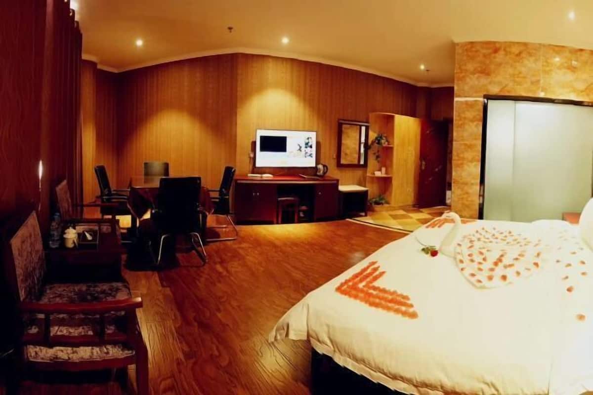 A hotel room with a wooden floor, a bed with a heart-shaped decoration, a desk with a computer, and a TV.