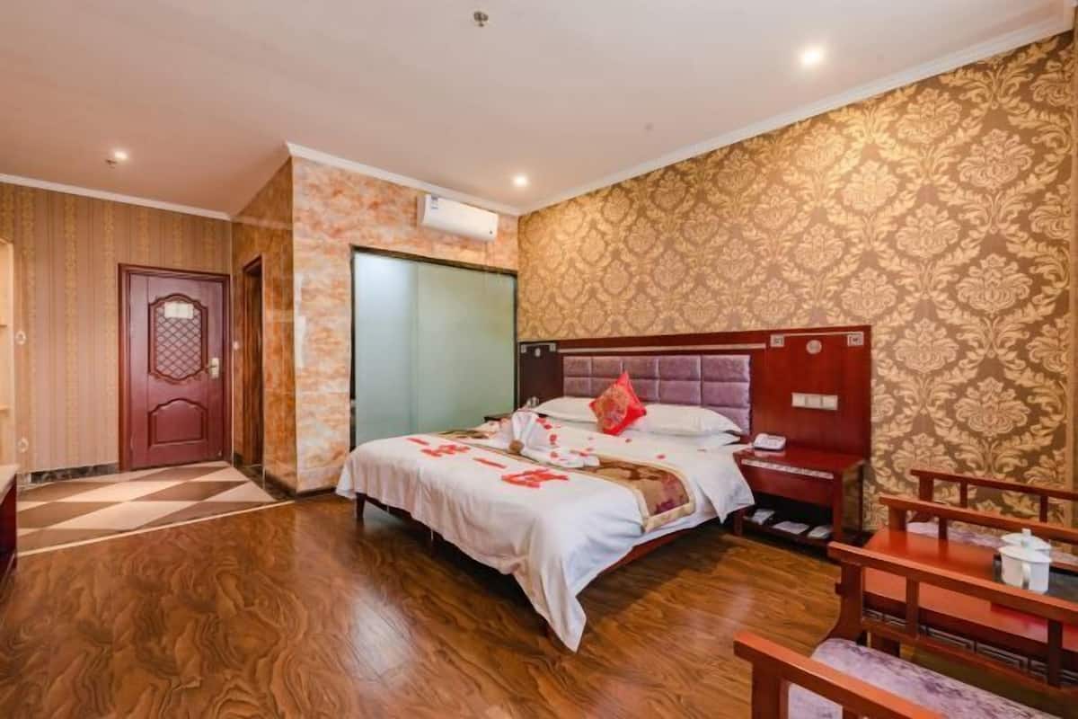 A hotel room with a wooden floor, a bed with a red pillow, a nightstand, and a wall with patterned wallpaper.