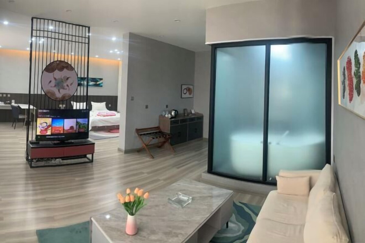 A modern hotel room with a large sliding glass door, a flat-screen TV, a sofa, and a coffee table with a vase of flowers.