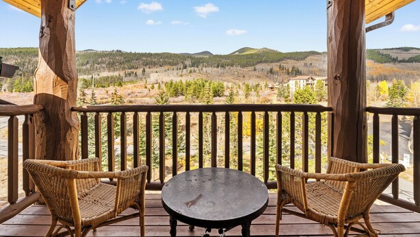 A wooden deck with two wicker chairs and a table, overlooking a forested landscape with hills and a building in the distance. - Hidden River Lodge #5998 by Summit County Mountain Retreats (Keystone)