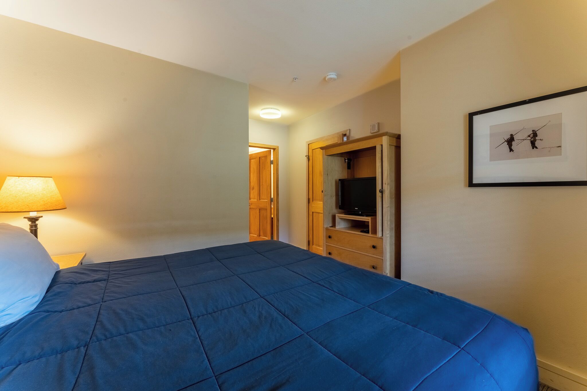 A hotel room with a large bed, a TV, a wooden wardrobe, and a framed picture on the wall.
