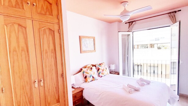 A bedroom with a large wooden wardrobe, a bed with white bedding, colorful pillows, a ceiling fan, and a view of the outside balcony.