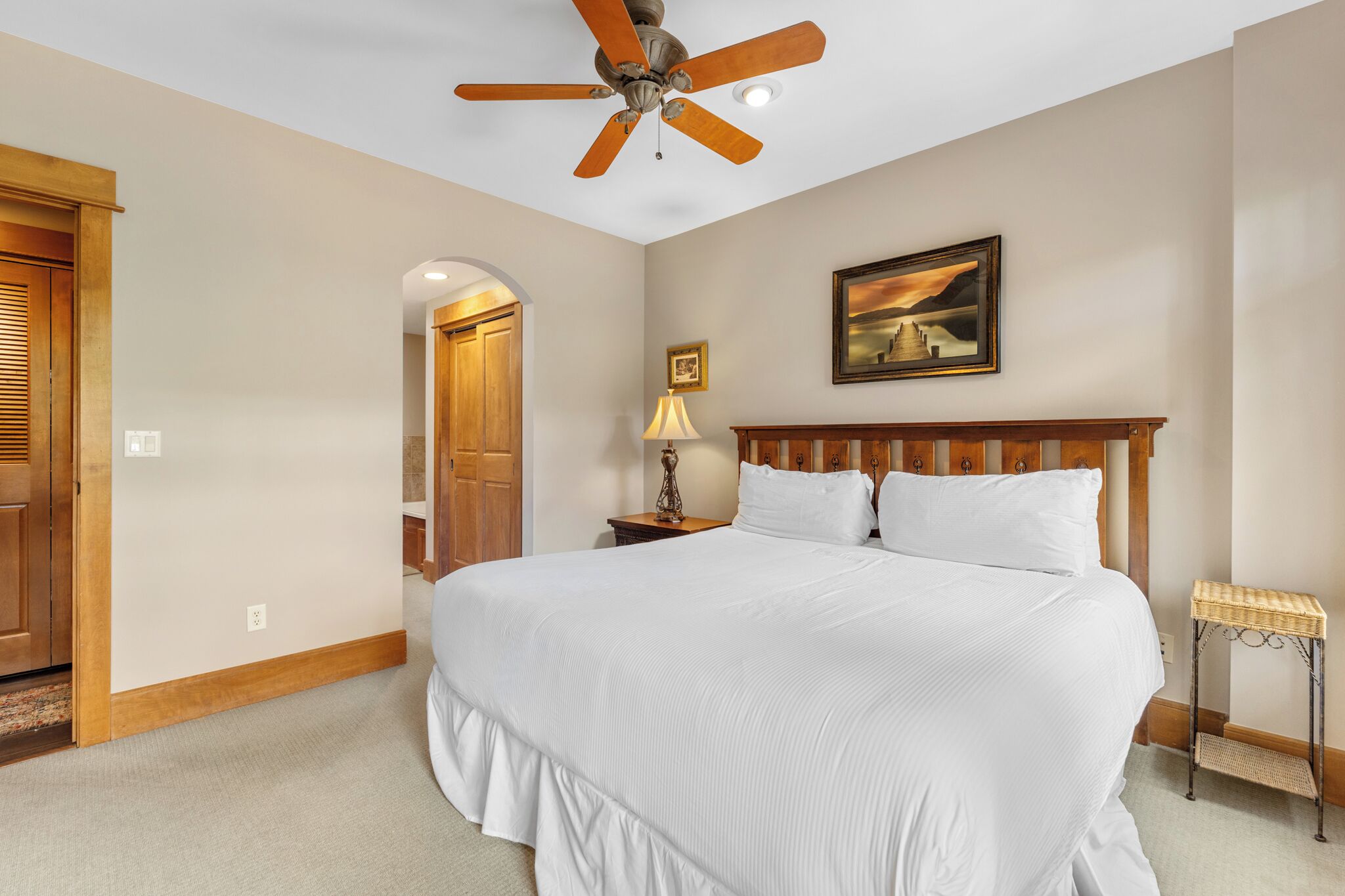 A bedroom with a large bed, a ceiling fan, a nightstand with a lamp, a framed picture on the wall, and an arched doorway.