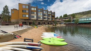 Um edifício moderno com fachada de pedra e várias janelas, uma praia arenosa com caiaques e pranchas de remo, e uma extensão de água calma.