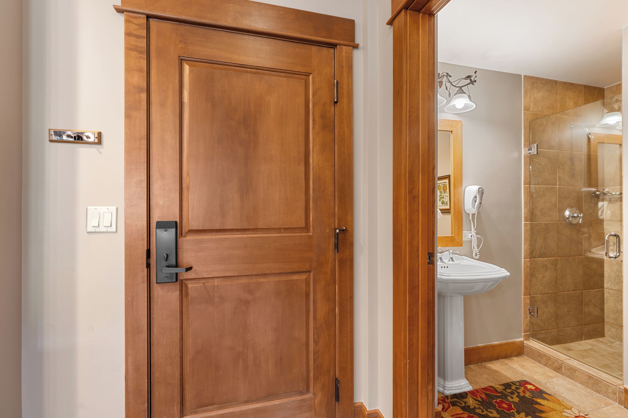 A wooden door with a modern handle, a white wall switch, and a bathroom with a sink and shower.