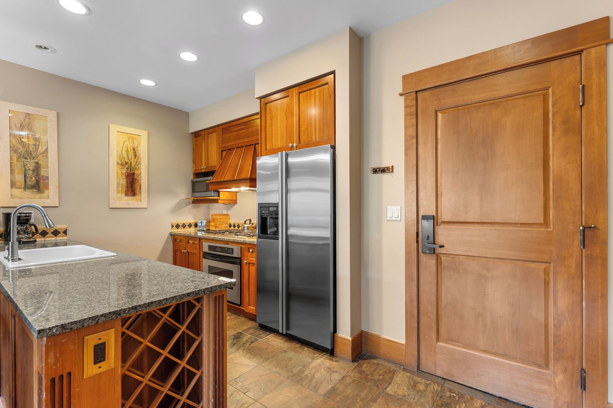 Una cocina con armarios de madera, un refrigerador de acero inoxidable, una encimera de granito y una estantería para vinos.